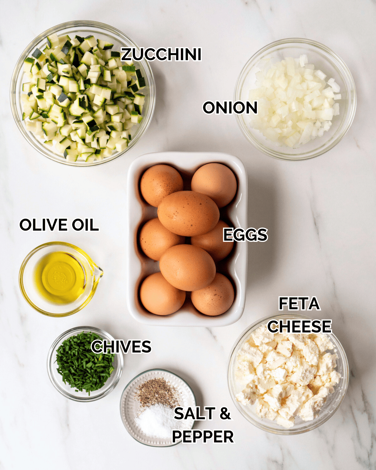 Ingredient shot of individual ingredients in clear bowls including eggs, feta cheese, salt and pepper, chives, olive oil, onion, and zucchini.